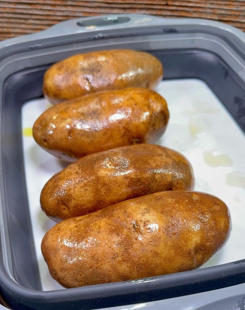 Four whole, unpeeled russet potatoes are placed in a rectangular, gray baking dish with a white interior, ready for baking. The potatoes have a slightly shiny appearance.