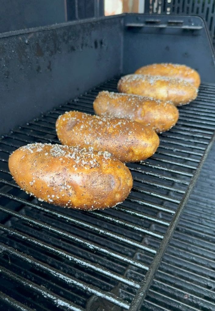 Five seasoned russet potatoes are lined up on the grates of a barbecue grill, being cooked. The potatoes appear to be coated with salt and spices, and the grill is partially open. Recipe for loaded baked potato salad