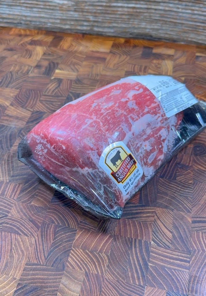 A vacuum-sealed cut of raw beef rests on a patterned brown cutting board. The packaging has a label that reads Certified Angus Beef and a smaller white label with product details.