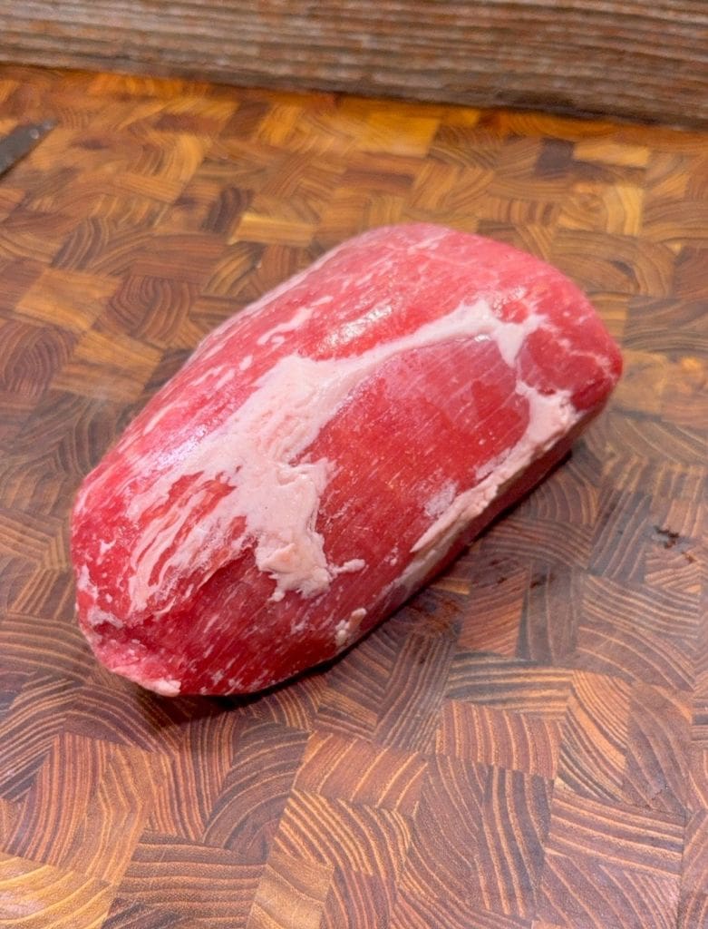 A raw, boneless cut of beef with some marbled fat rests on a wooden cutting board with a patterned surface.