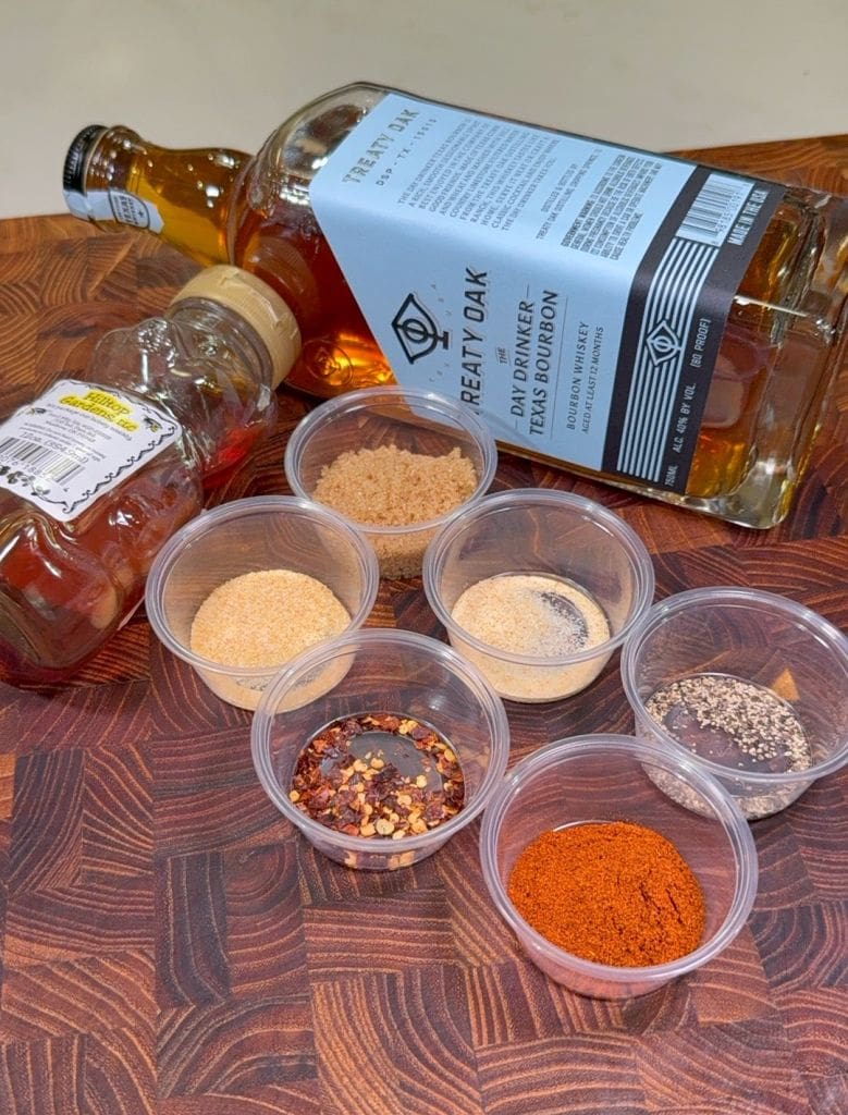 A bottle of Real Dry Oak Texas Bourbon and a small bottle of honey lie on a wooden surface next to small plastic containers holding various spices and seasonings, including brown sugar, chili flakes, and ground paprika.  Ingredients for the marinade of bourbon beef jerky 