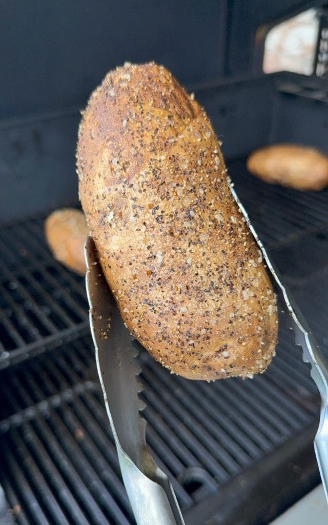 A baked potato coated with pepper and seasoning is held by metal tongs in front of a grill, with other seasoned potatoes visible in the background.