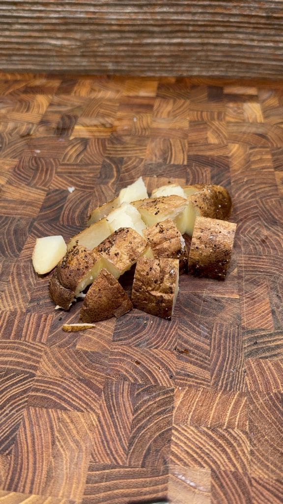 Diced, cooked potato pieces with skin and black pepper on a wooden cutting board.