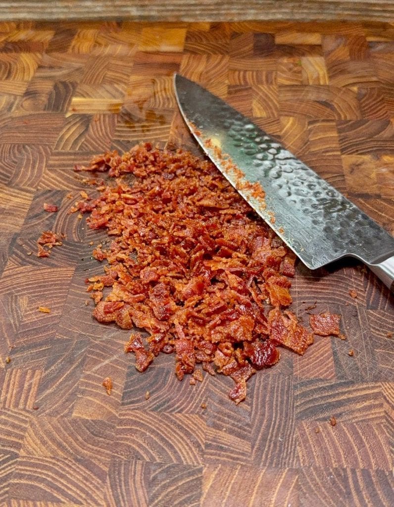 A chef’s knife rests beside a small pile of finely chopped crispy bacon pieces on a wooden cutting board with a mosaic pattern.
