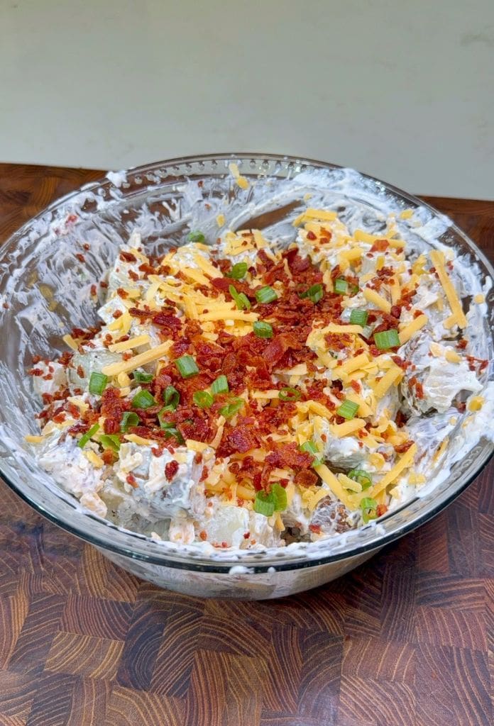 A glass bowl filled with creamy potato salad, topped with shredded cheddar cheese, chopped green onions, and crumbled bacon on a wooden surface.