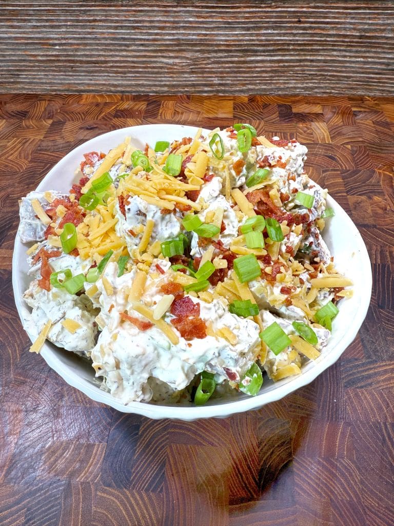 A bowl of loaded potato salad topped with shredded cheddar cheese, chopped green onions, and bacon bits, set on a wooden surface.
