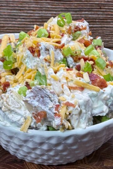 A white bowl filled with loaded potato salad, topped with shredded cheese, bacon bits, and chopped green onions, sitting on a wooden surface with a rustic wooden background.