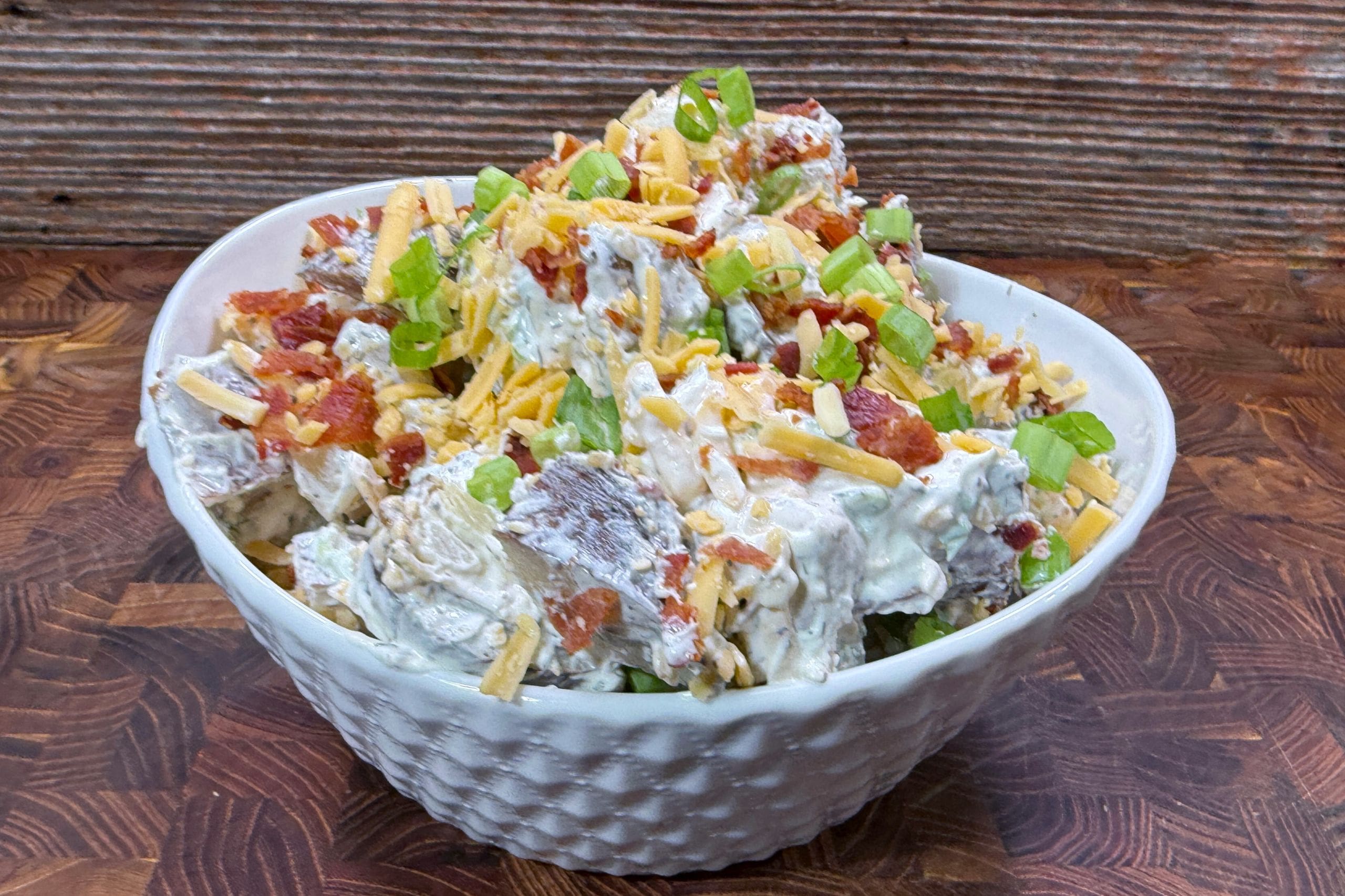 A white bowl filled with loaded potato salad, topped with shredded cheese, bacon bits, and chopped green onions, sitting on a wooden surface with a rustic wooden background.