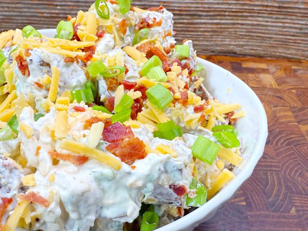 A bowl of loaded potato salad topped with shredded cheddar cheese, chopped green onions, and pieces of crispy bacon, set on a wooden surface.