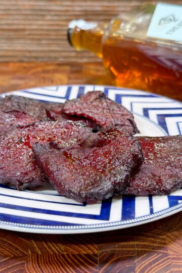 A plate with several pieces of dark, seasoned beef jerky sits on a blue and white patterned dish. In the background, a bottle of whiskey lies on its side on a wooden surface.
