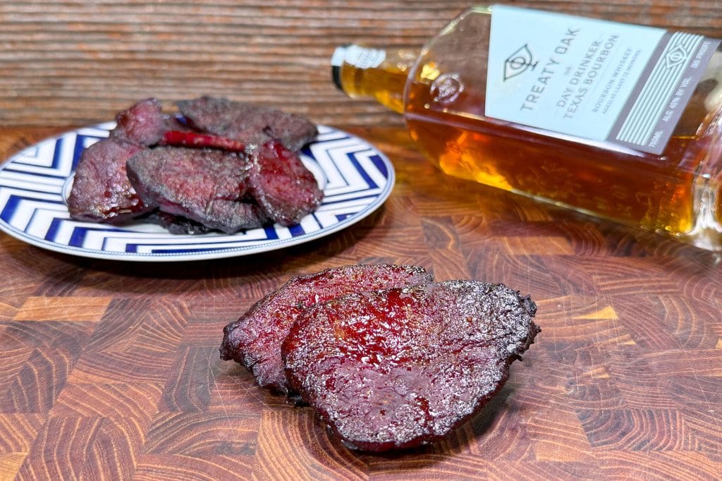 Two pieces of dark, grilled meat are on a wooden surface, with more pieces on a patterned plate in the background. A tipped bottle of Treaty Oak bourbon lies beside the plate.