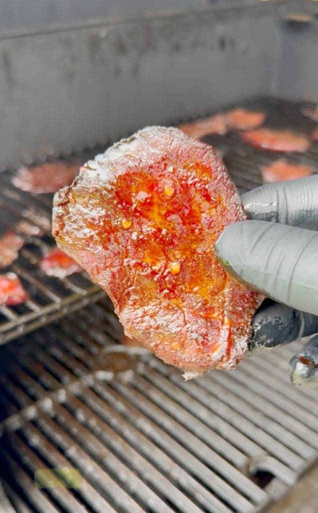 A gloved hand holds a shiny, glazed slice of smoked meat in front of a grill with more pieces cooking in the background. The meat appears caramelized and crispy around the edges.