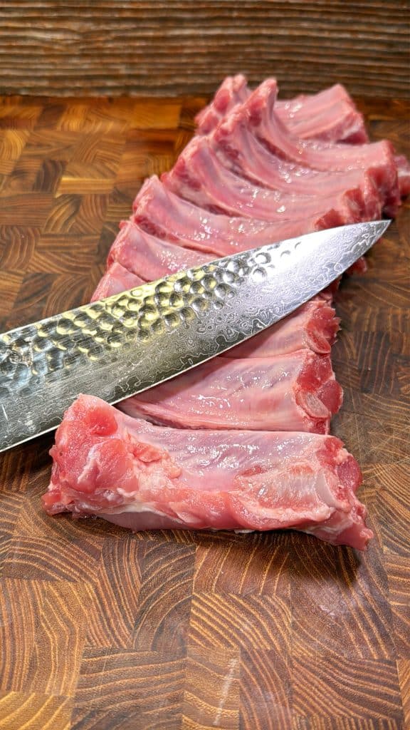 A close-up of raw pork ribs being sliced with a textured chef’s knife on a patterned wooden cutting board.