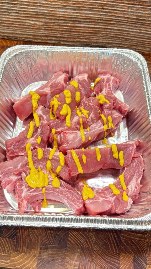 Raw pork ribs in a rectangular aluminum tray, with yellow mustard drizzled over the top, sitting on a wood surface.
