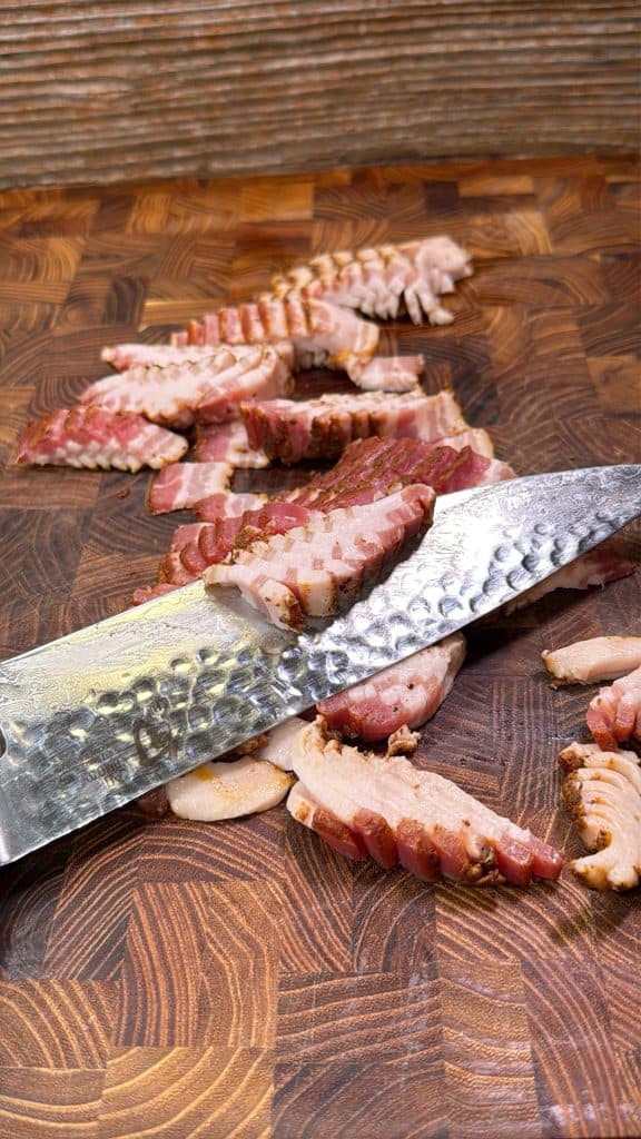 Slices of cooked, seasoned meat are spread on a wooden cutting board. A large chefs knife with a hammered blade lies across the meat, with a textured wooden background in the distance.