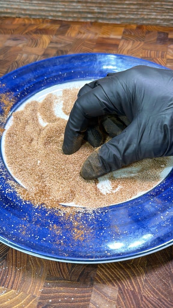 A hand in a black glove mixes a brown spice blend on a blue and white plate, placed on a wooden surface.