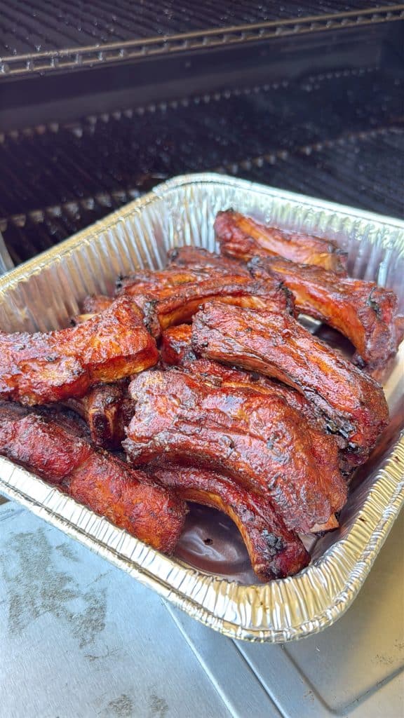 A disposable aluminum tray filled with freshly grilled, charred pork ribs sits on a metal surface in front of a barbecue grill. The Dr Pepper Party ribs are cooked and have a caramelized, crispy exterior.