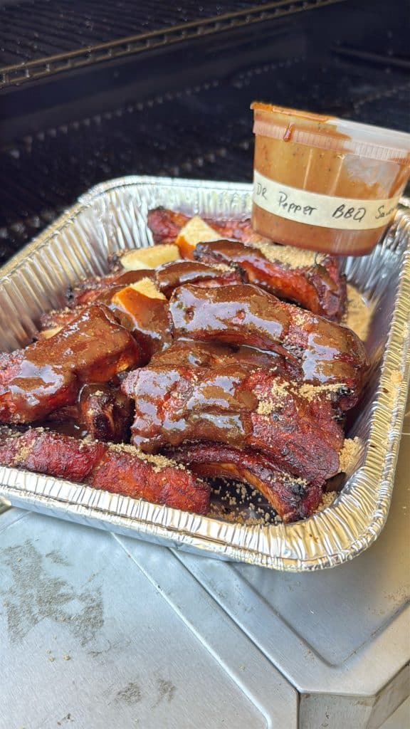 A foil tray filled with glazed, saucy barbecue ribs sits on a grill, with a container labeled Dr Pepper BBQ Sauce resting at the corner of the tray.
