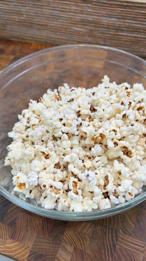 A clear glass bowl filled with freshly popped popcorn sits on a wooden surface. The popcorn is lightly browned in some spots, suggesting it is homemade.