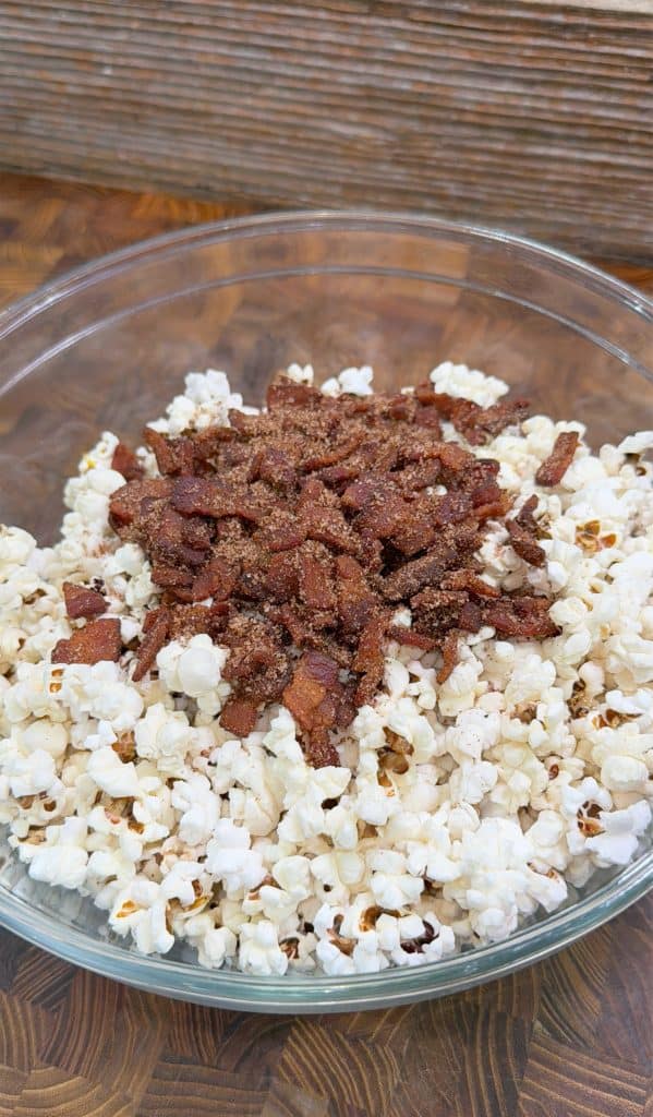 A clear glass bowl filled with popcorn, topped with crumbled, seasoned bacon pieces, sits on a wooden surface.