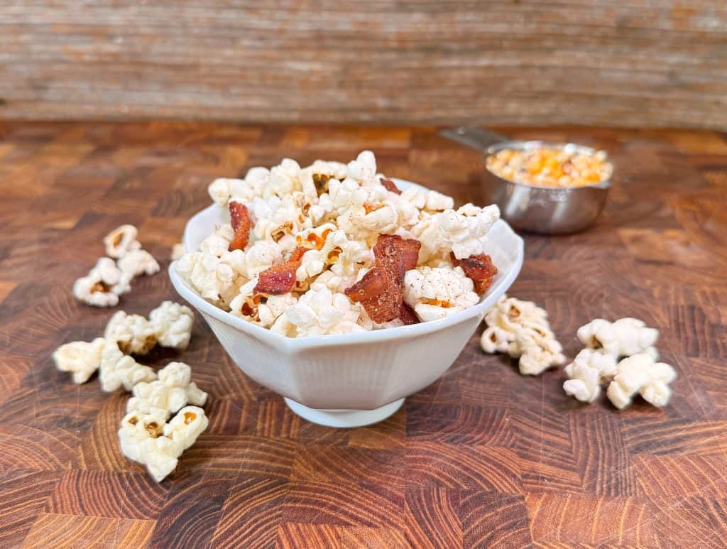 A white bowl filled with popcorn and bacon pieces sits on a wooden surface, surrounded by loose popcorn. In the background, a metal measuring cup holds unpopped corn kernels. Recipe is for candied bacon popcorn