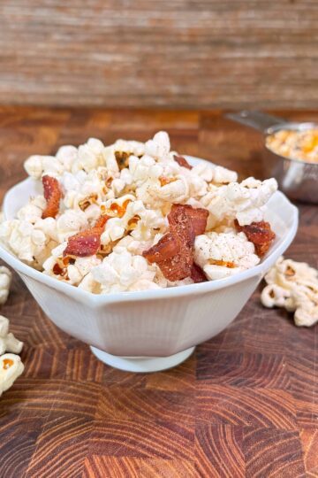 A white bowl filled with popcorn and bacon pieces sits on a wooden surface, surrounded by loose popcorn. In the background, a metal measuring cup holds unpopped corn kernels.