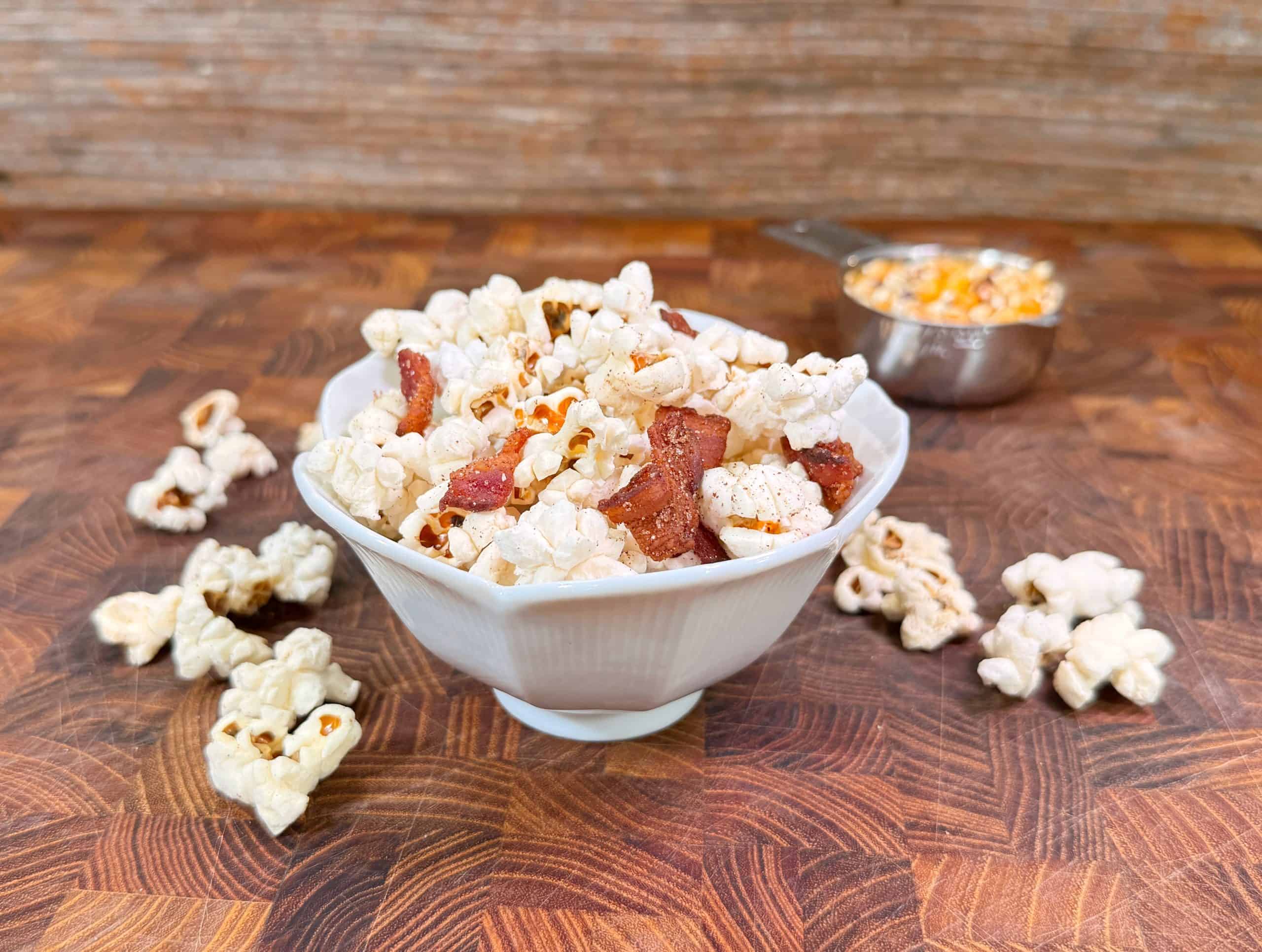 A white bowl filled with popcorn and bacon pieces sits on a wooden surface, surrounded by loose popcorn. In the background, a metal measuring cup holds unpopped corn kernels.