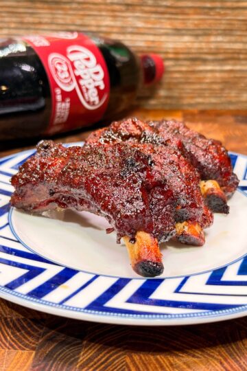 A plate with grilled ribs sits on a blue and white zigzag-patterned dish, with a bottle of Dr Pepper soda lying on its side in the background on a wooden surface.