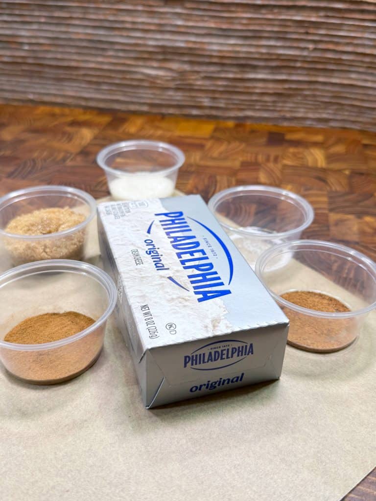 A block of Philadelphia original cream cheese is surrounded by six small plastic cups containing various ingredients, including brown sugar, white sugar, and spices, all placed on a wooden surface.