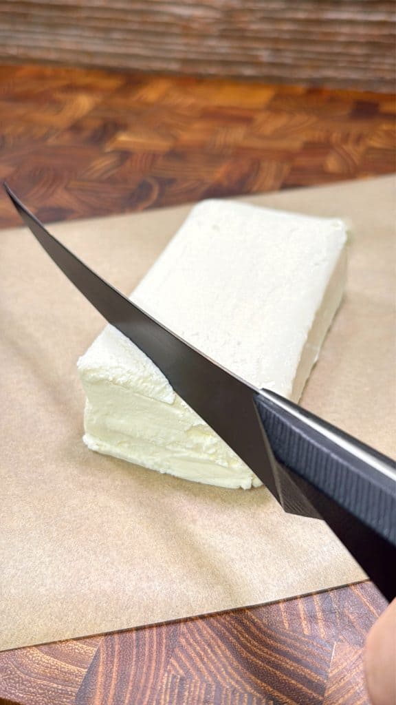 A close-up of a hand holding a knife, slicing through a block of cream cheese on brown parchment paper atop a wooden surface.