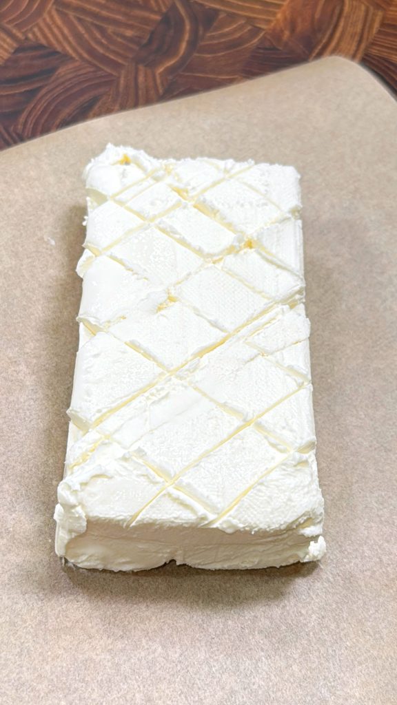 A rectangular block of cream cheese with a crisscross pattern scored on top, resting on a sheet of parchment paper.