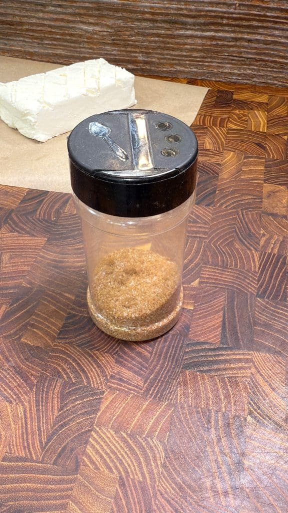 A glass sugar dispenser with a metal lid, containing brown sugar, sits on a patterned wooden surface. In the background, there is a block of white cheese on parchment paper.