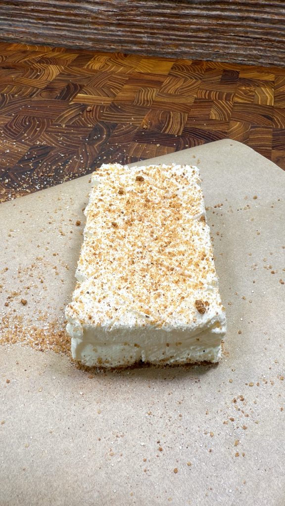 A rectangular slice of creamy dessert with a crumb topping sits on a sheet of brown parchment paper, against a wooden background. Crumbs are scattered around the dessert.