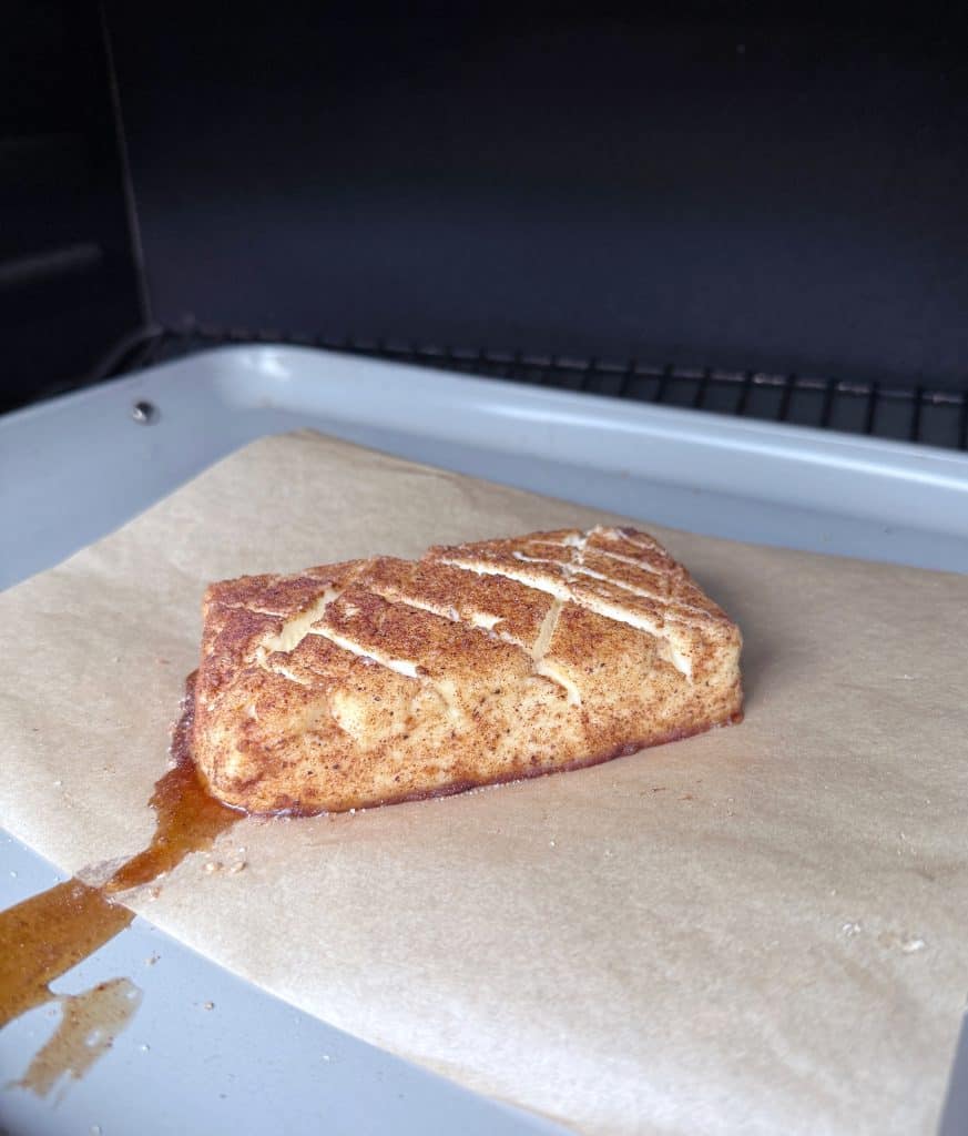 A single frosted pastry with cinnamon sugar and a drizzle of icing sits on parchment paper on a baking tray, with a bit of filling oozing from one side.