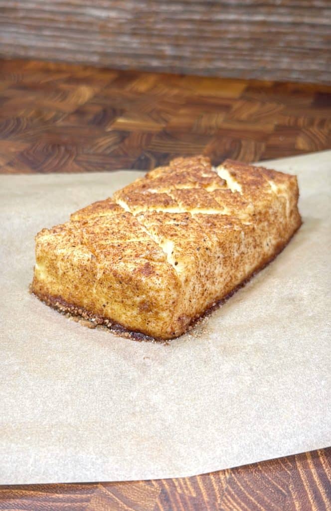 A rectangular loaf of smoked cinnamon roll cream cheese with a golden-brown crust and cinnamon sugar coating sits on parchment paper atop a wooden surface.