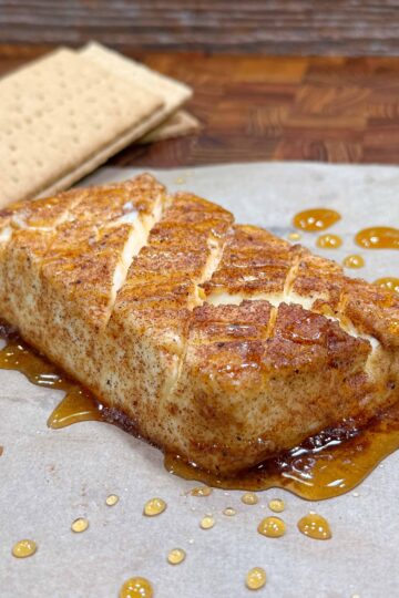 A block of seasoned, grilled cream cheese with a caramelized glaze, drizzled with honey and served on parchment paper, with two graham crackers in the background.