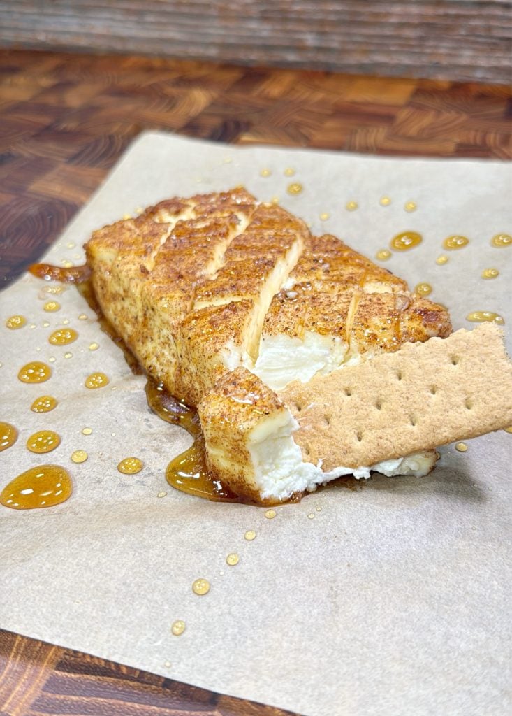 A graham cracker dipped into a block of cream cheese coated with cinnamon and sugar, drizzled with honey, served on parchment paper on a wooden surface.