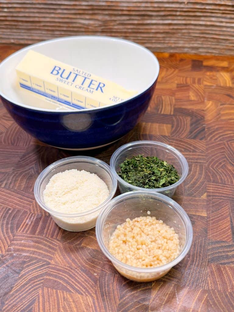 A blue and white bowl with a stick of butter sits behind three small containers holding grated Parmesan cheese, chopped herbs, and minced garlic on a wooden surface.