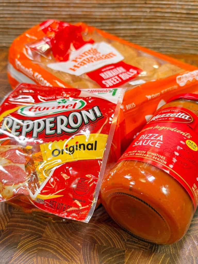 A package of sandwich buns, a bag of Hormel original pepperoni, and a jar of Mezzetta pizza sauce on a wooden surface.