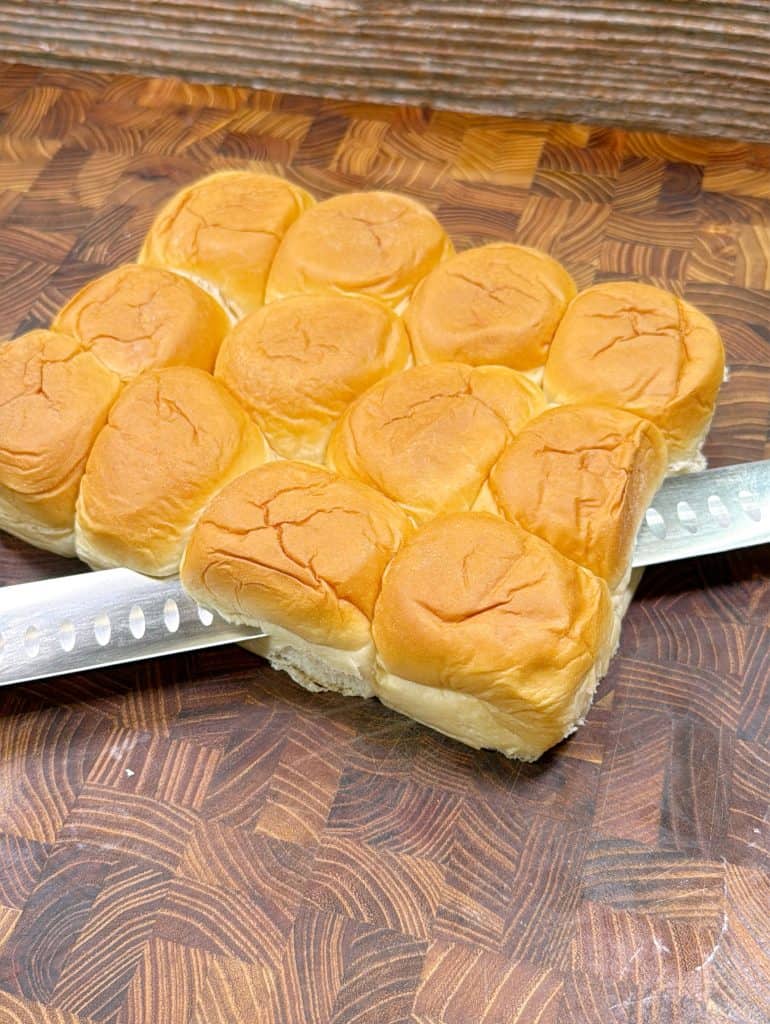 A serrated knife is slicing through a cluster of soft, golden brown dinner rolls on a patterned wooden cutting board.