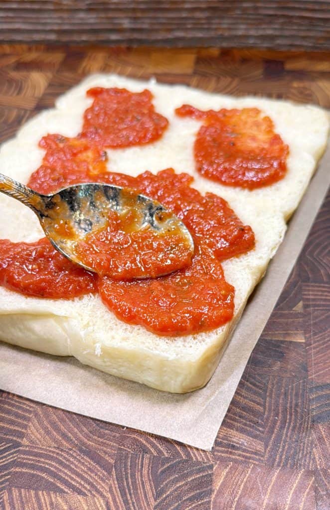 A spoon spreads tomato sauce over a thick, rectangular piece of raw pizza dough on parchment paper, set on a wooden surface.