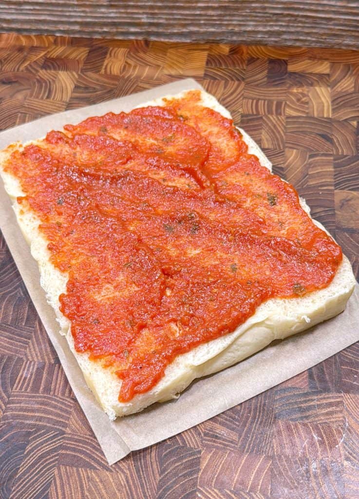 A thick slice of focaccia bread topped with a generous layer of tomato sauce sits on a piece of parchment paper on a wooden cutting board.