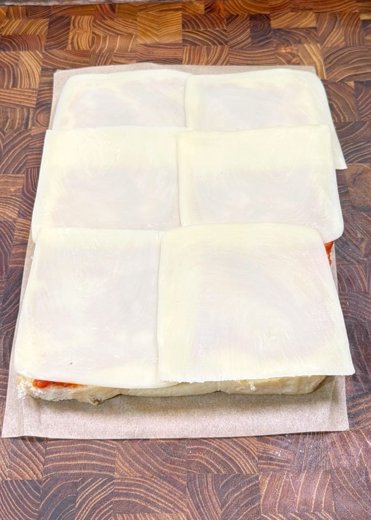 Six slices of white cheese are layered on top of stacked bread and a visible layer of sauce, sitting on a wooden cutting board.