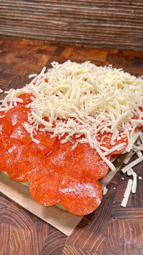 Rectangular piece of pizza dough on a wooden surface, half covered with sliced pepperoni and the other half topped with shredded mozzarella cheese.