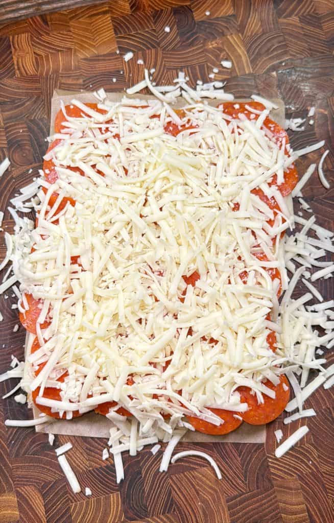 Rectangular sheet of dough topped with pepperoni slices and a generous layer of shredded mozzarella cheese, resting on a wooden cutting board.