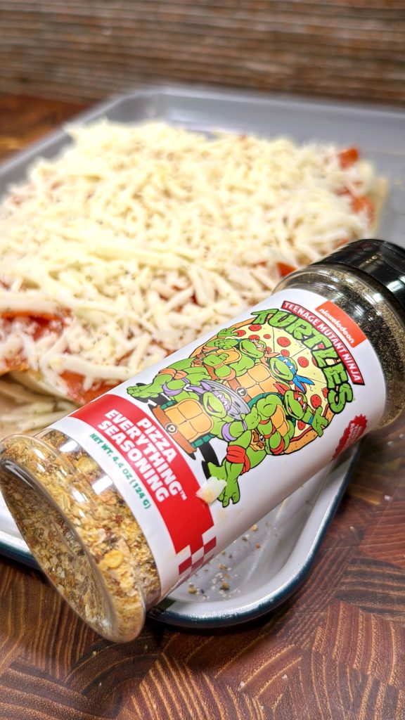 A bottle of Teenage Mutant Ninja Turtles Pizza Seasoning lies next to an uncooked pizza covered with shredded cheese on a baking tray.