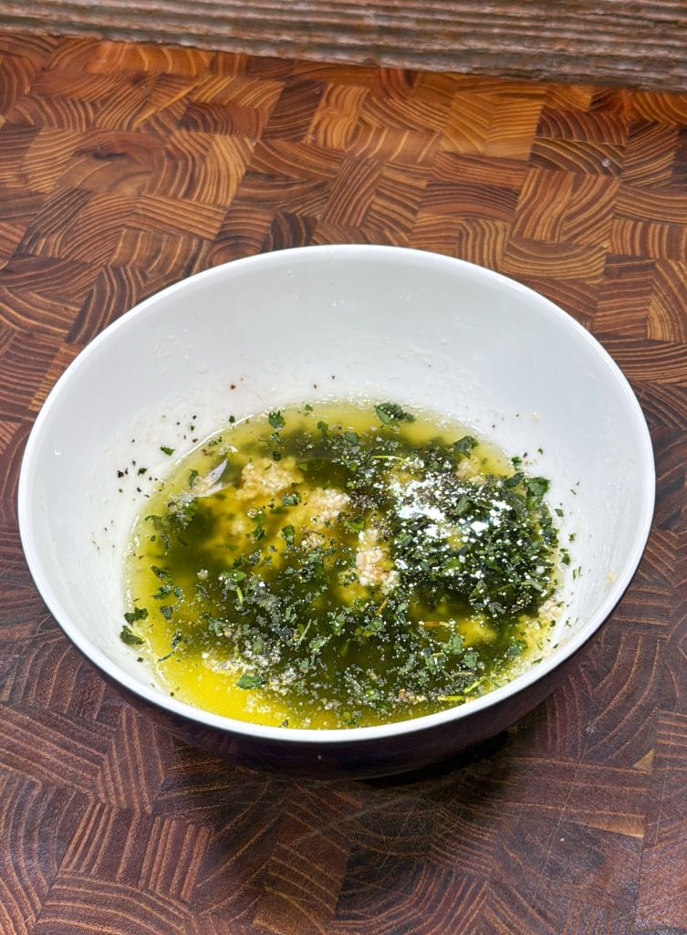 A white bowl on a patterned wooden surface contains melted butter mixed with chopped herbs and grated cheese.