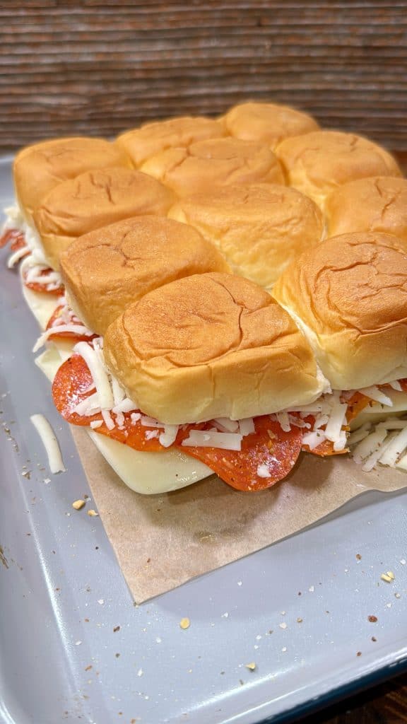 A close-up of a tray of unbaked slider sandwiches filled with pepperoni and shredded cheese on a sheet of parchment paper. The top buns are slightly browned and soft.