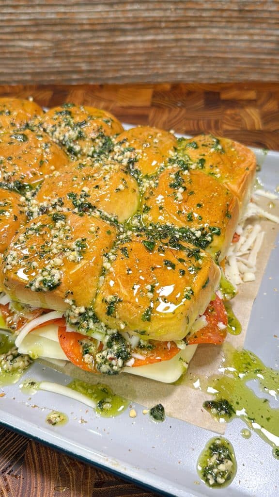 Pizza Sliders topped with a green herb and cheese mixture, filled with layers of cheese and pepperoni, are arranged on a tray, ready to be baked or served.