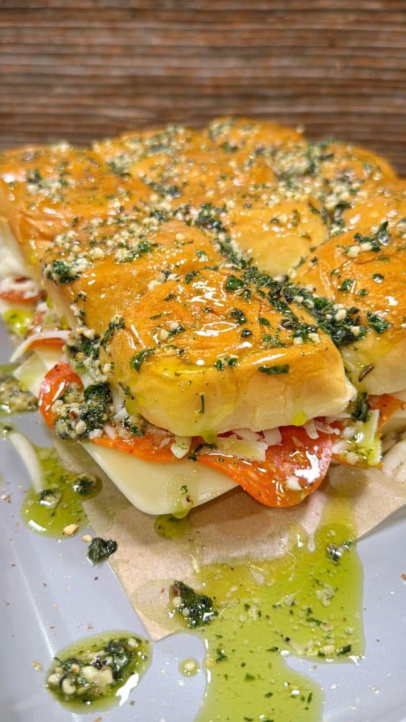 A close-up of a row of baked Italian-style slider sandwiches filled with meats, cheese, and shredded lettuce, topped with a shiny, herb-garlic butter glaze, with extra seasoning and oil drizzled around them.